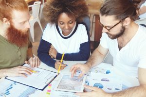 Youth, education and technology concept. Group of three young smart and intelligent future marketing specialists working together on graduation project, analyzing graphs and charts, using touch pad